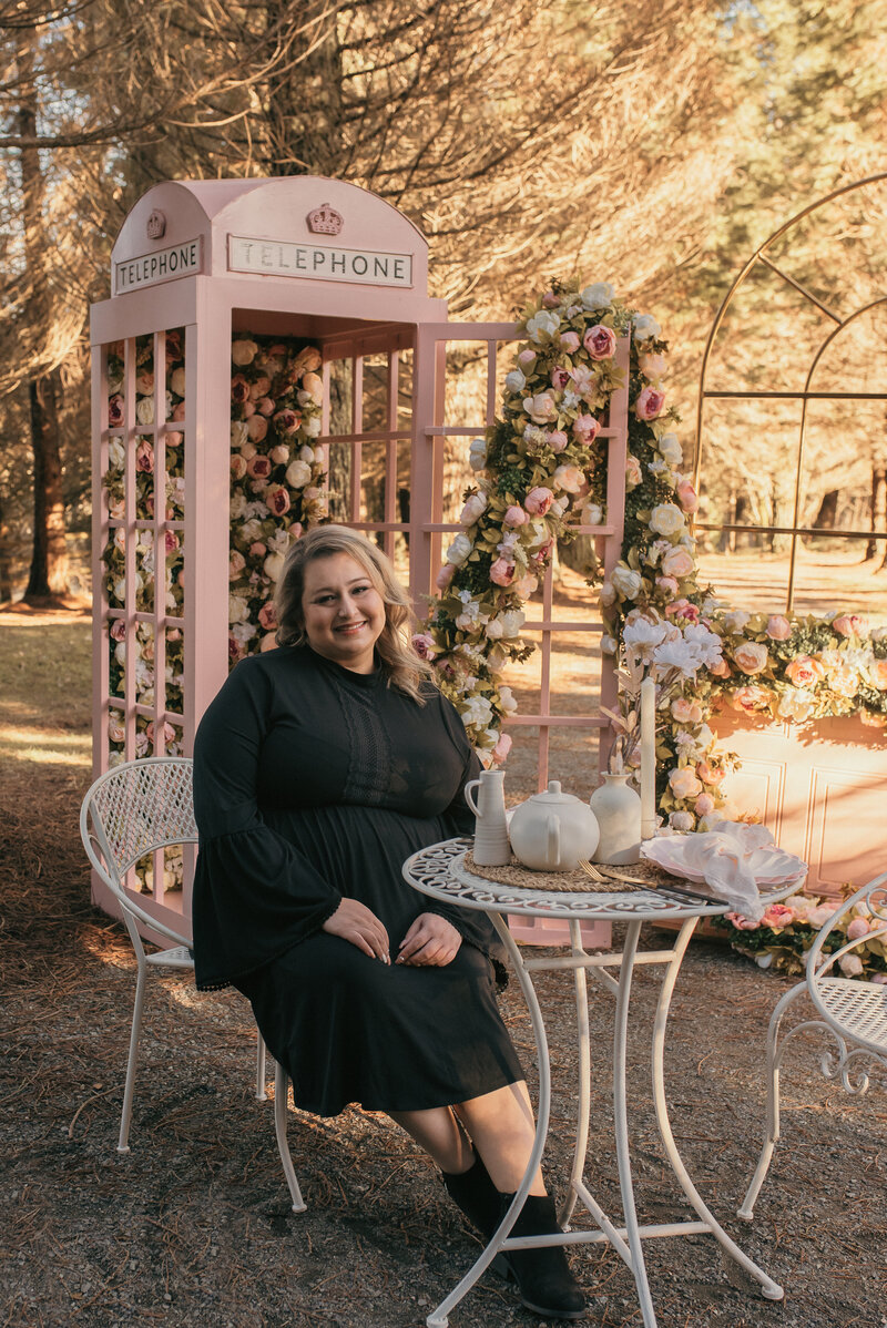 wedding planner sitting at a small white table next to a white teapit and a floral filled pink telephone booth