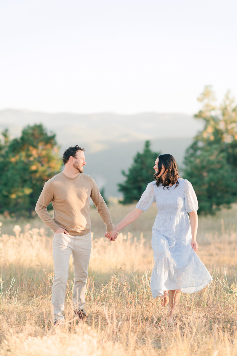 Melanie and Tyler hold hands and walk through a field with mountains in the background.