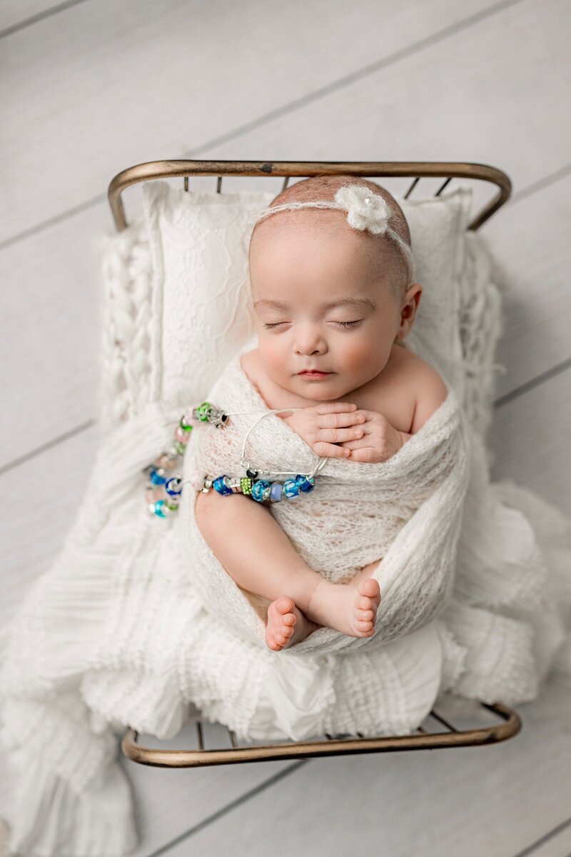 Newborn baby girl is wrapped in a white wrap holding beads from her time in the NICU laying on a bed with white layers in a Charlotte Area Studio