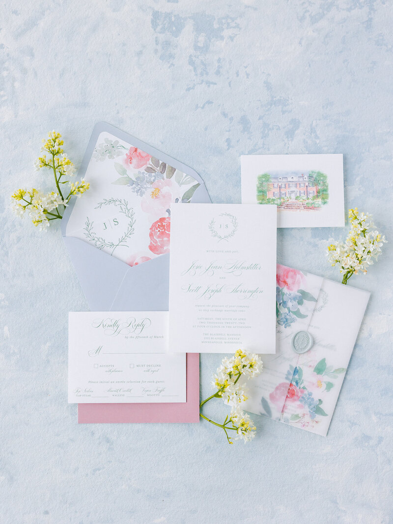 Flat lay of a colorful floral themed wedding invitation suite on a light blue patterned background