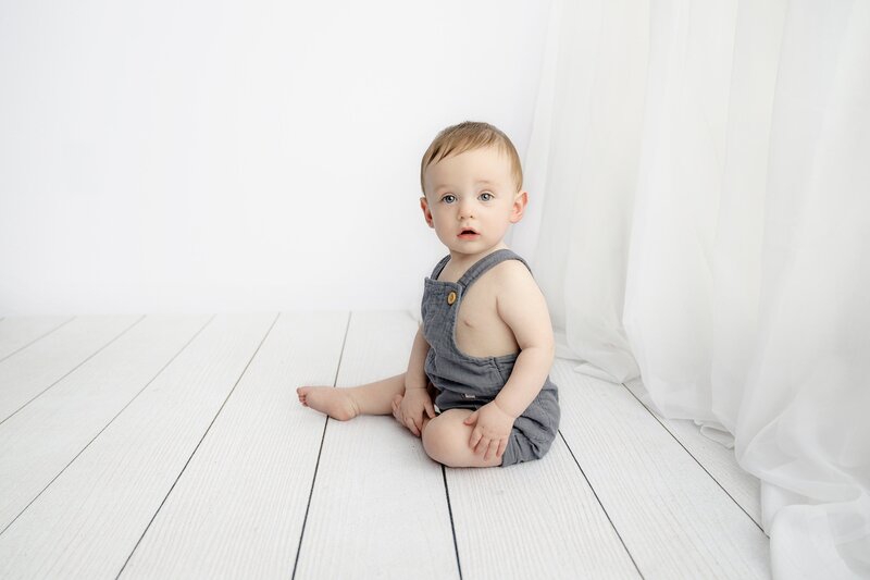 boy sits in blue grey overalls in a white background