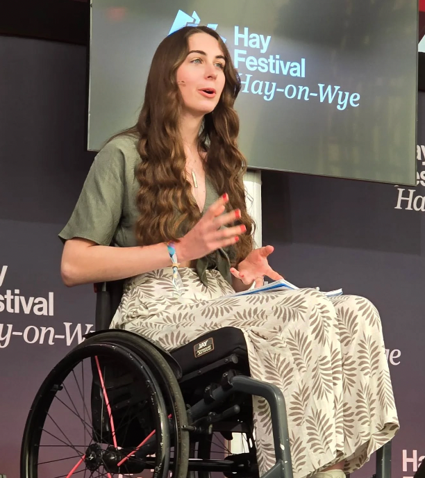 Bethany Handley speaking on stage at Hay Festival, seated in a wheelchair with a notepad in hand.