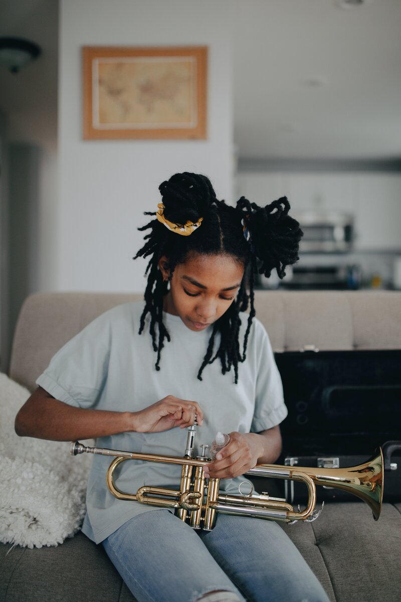 A teen with long braids pulled up into two buns sits on a couch, a trumpet in her lap. She is cleaning the valves with a focused, downturned expression.