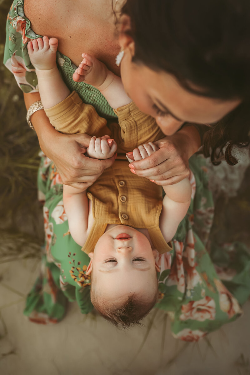 Little boy in mustard yellow overalls lays on mom's lap upside down  while mom in summer floral dress hold his tiny fingers and smiles at him