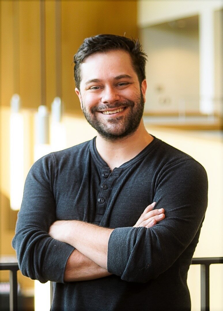 A cheerful man with stubble, wearing a black cardigan over a dark shirt, arms crossed