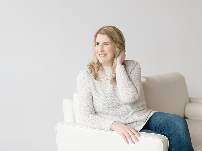 Photo of Seattle photographer, Shaunae Teske, a woman with blond hair wearing a cozy knit sweater sitting on a plush white couch in a photo studio looking off to the side and smiling