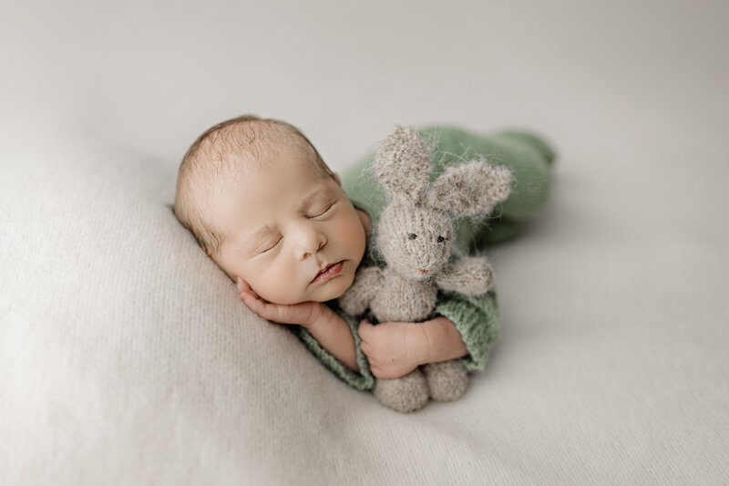 A sleeping newborn baby in a green onesie is lying on their side, holding a knitted stuffed rabbit.