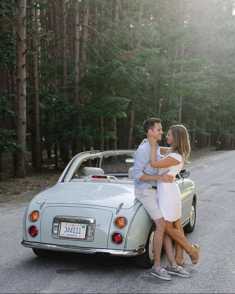 Couple leaning on a classic car, holding eachother and smiling during their engagement shoot  with pine tress in the background