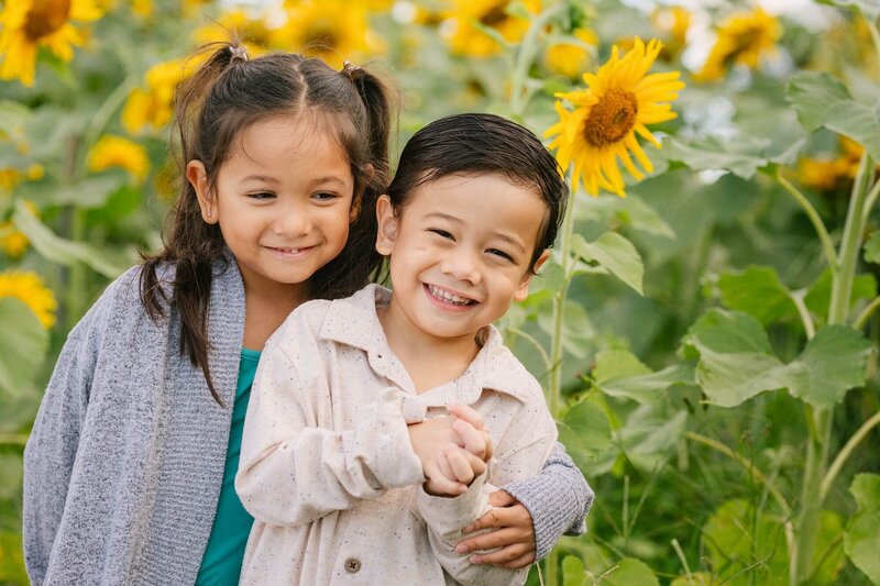 A little boy and girl hug smiling with sunflowers in the background.