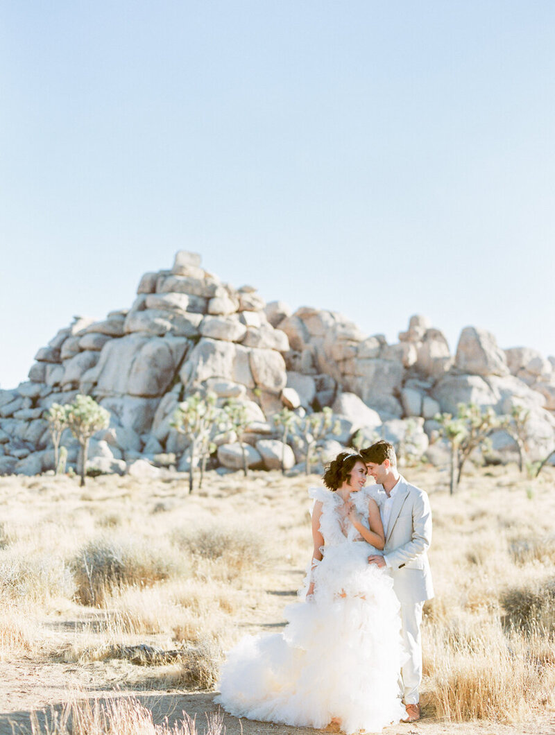 bride  in a poofy wedding dress and groom in a tan suit with their foreheads together with a rock formation and palm trees behind them in  Joshua Tree National Park