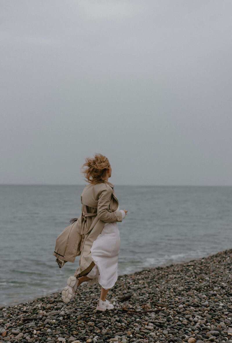 Frau im Trenchcoat und Kleid, am Strand lang laufend, der Blick nach vorne, der Wind im Haar: Ziele setzen und entschlossen voranschreiten