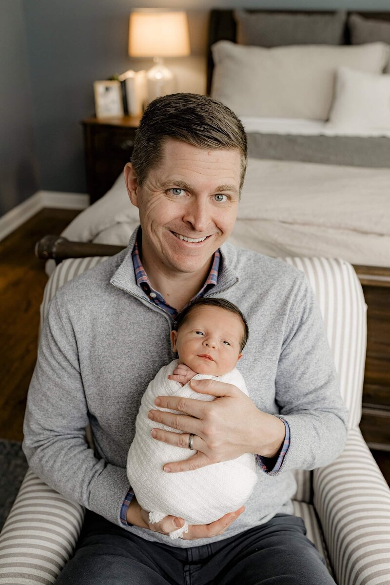 father holds his newborn son wrapped in white sitting in their master bedroom during their lifestyle newborn session in their Charlotte Area home