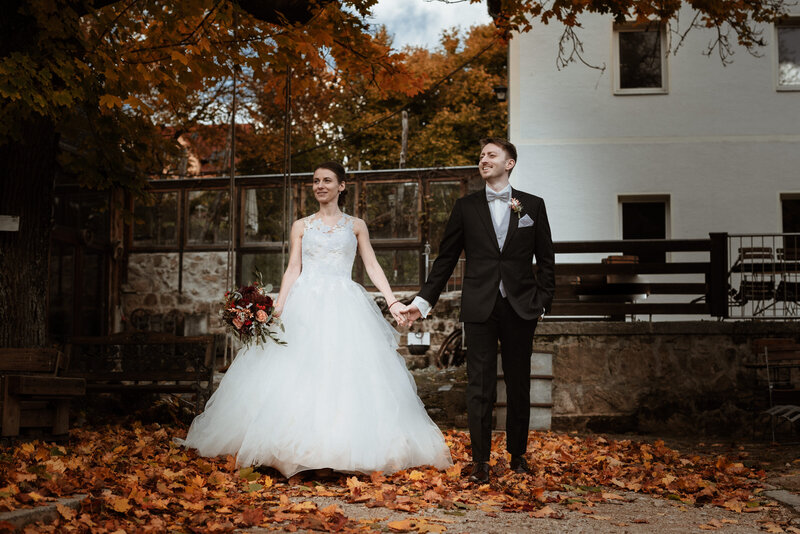 Hochzeitsfoto eines Brautpaares nebeneinander stehend inmitten eines Hofes voller buntem Herbstlaub.