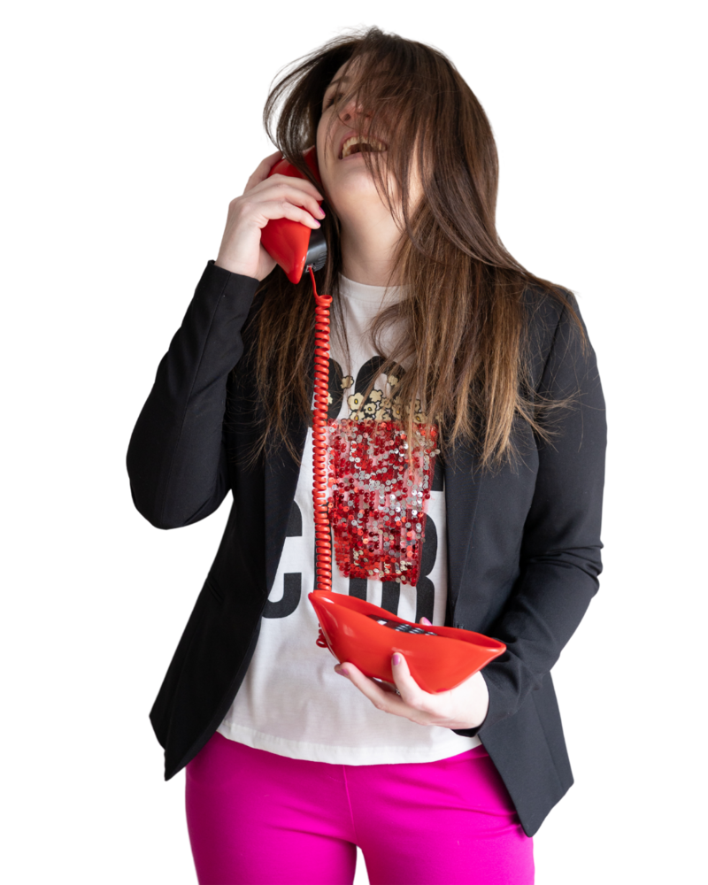 Emily wearing hot pink pants, a white shirt that says popcorn with a glittery image of popcorn on it and a black blazer. She is holding a red lip shaped phone up to her ear and smiling with her hair covering her face