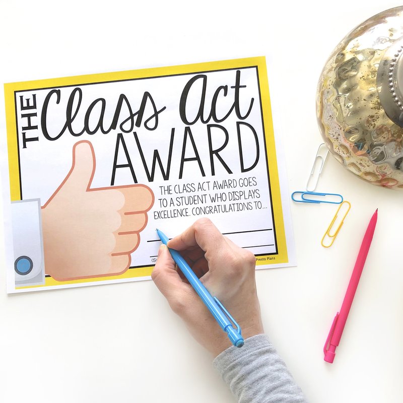 A teacher writing on an end of the year  student award where each award relates to a different idiom.  In this one, the award is the class act award and it goes to a student who displays excellence.  The award is beside a pencil, paperclips, and a decorative jar.