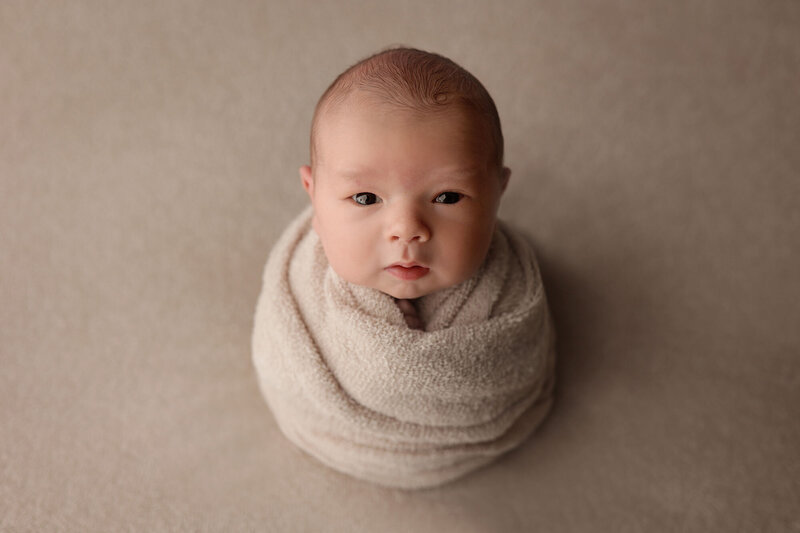 awake baby swaddled in a beige wrap posed by an experienced newborn photographer