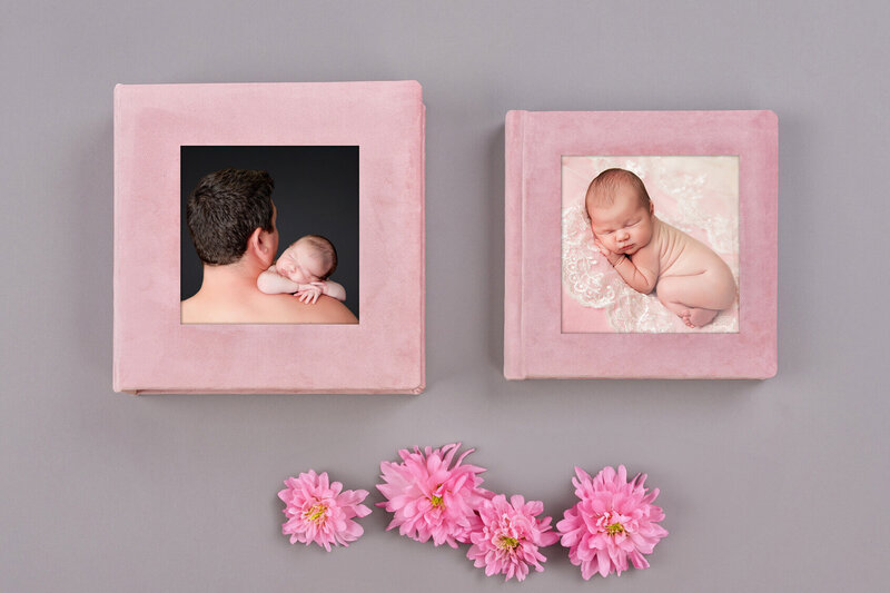Two pink photo albums featuring baby portraits, perfect for showcasing newborn photography in Jacksonville, are placed side by side on a gray surface. Three pink flowers are arranged underneath the albums.