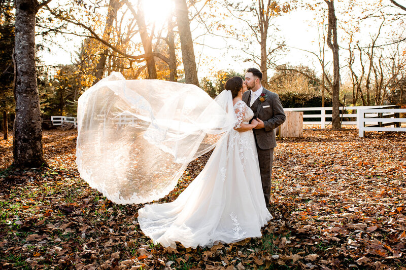 Dramatic photo of brides cathedral veil on the wind while kissing her husband outside in the fall leaves at golden hour