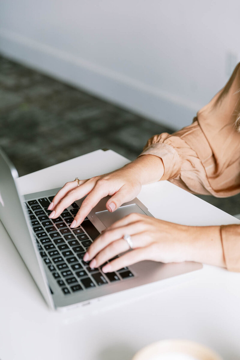 Hands typing on laptop keyboard with woman in beige clothing, representing digital engagement and online assessment taking.