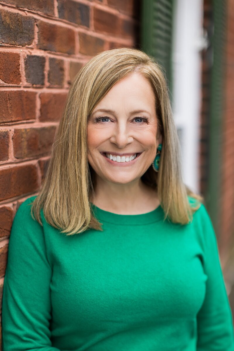 A blonde woman in a green top smiling against a brick wall