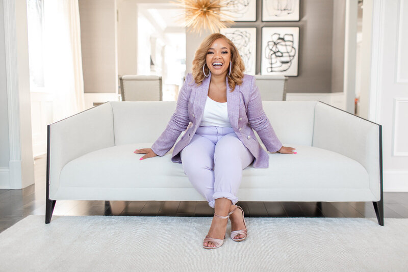 woman in a purple suit smiling into camera while sitting on a white couch