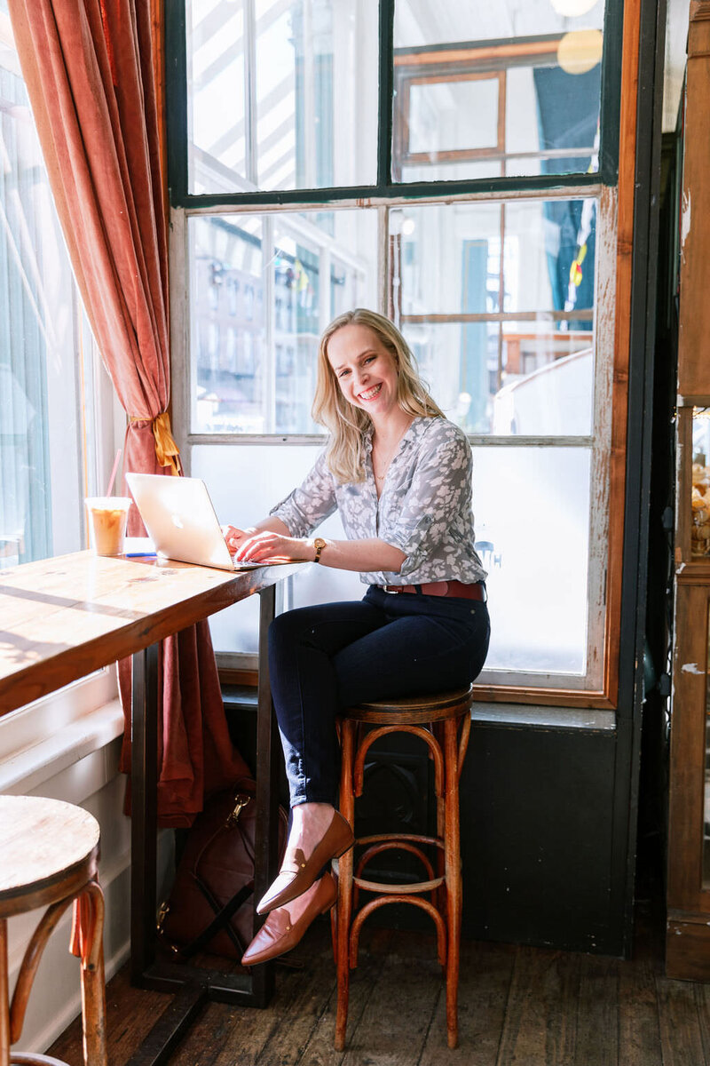 Digital Grace Design owner and Showit website designer, Sarah Blodgett, sits in a French inspired cafe on a stool in front of a window and smiles while working on her laptop with an iced coffee