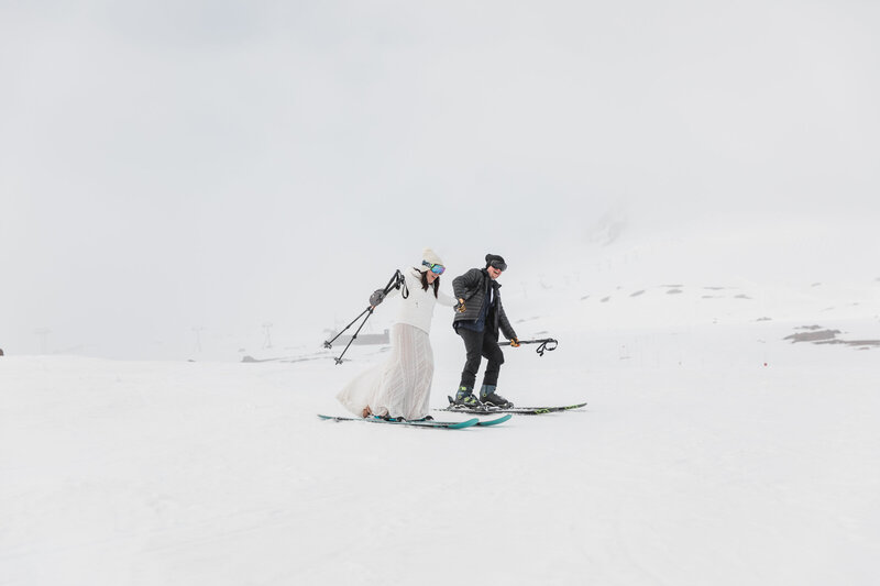 A bride on skis wearing a white dress  and Patagonia down jacket holds her groom in a black tux and black Patagonia own jacket's hand, as they ski down the slopes at Timberline Resort after saying their vows at their intimate Silcox Hut wedding on Mt. Hood  in Oregon. | Erica Swantek Photography