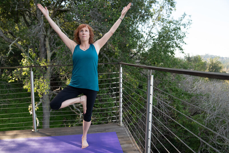 Woman outside on a deck standing on a yoga mat in a simple one legged yoga pose