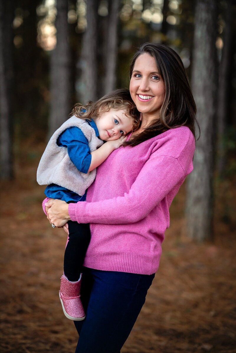A mom in a pink sweater is holding her young daughter as they stand in a forest.