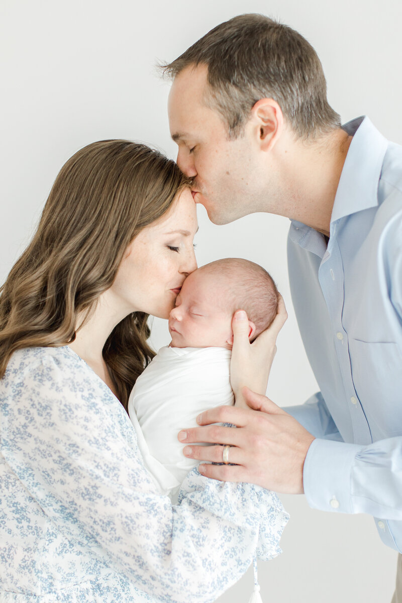 New mother kisses her sleeping baby's forehead as her husband kisses hers during newborn photography session
