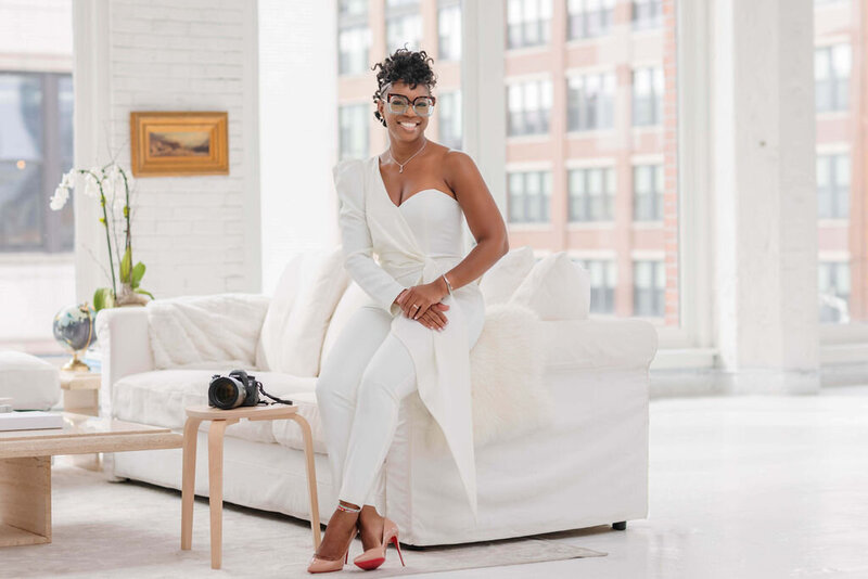 woman in a white suit sitting on the edge of a white couch next to a camera