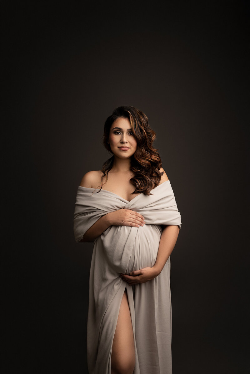 Woman poses for a fine art maternity photos at New Jersey portrait studio. Woman is artfully draped in a linen-coloured fabric. Her shoulders are bare and the fabric is ruched on her upper arm. One hand is above her belly and one other. She has a closed-mouth smile with her curled brown hair draping over her shoulder. Captured by best maternity photographer in New Jersey Katie Marshall.