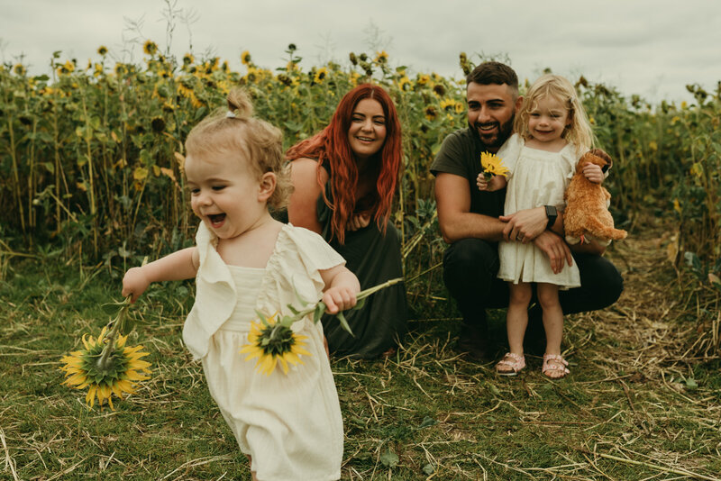 A mother and father holding their little girl in a field of sunflowers while there other little girl is walking out of shot