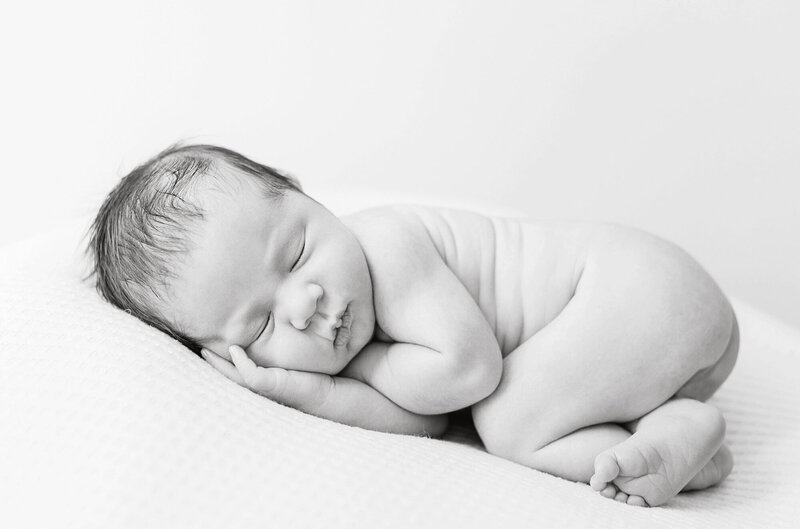A black and whitel photo of a newborn baby curled up and sleeping in a cute pose during his newborn photography session