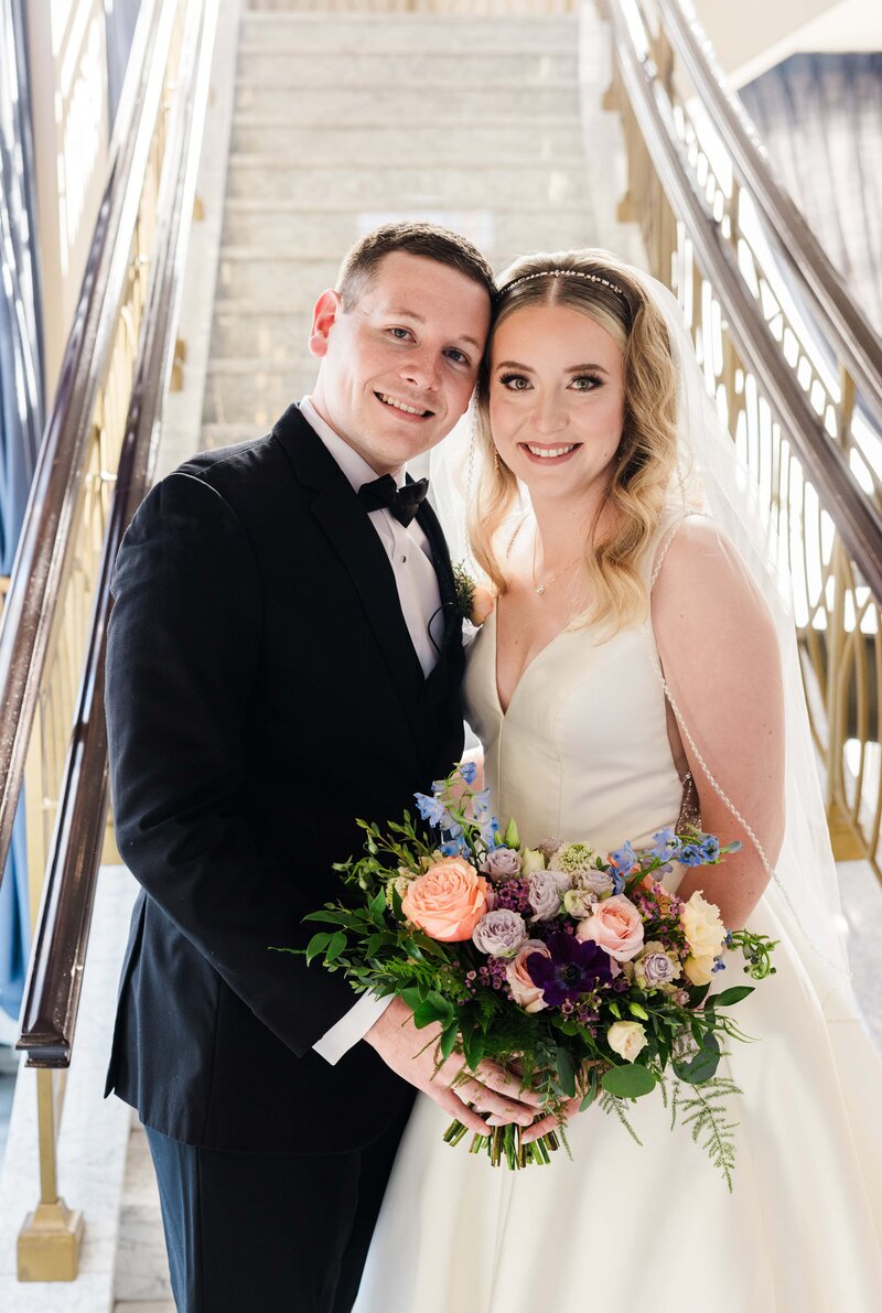 A bride and groom smiling on a staircase at an event in Davenport, the bride holding a bouquet with pastel flowers, both dressed in formal wedding attire.