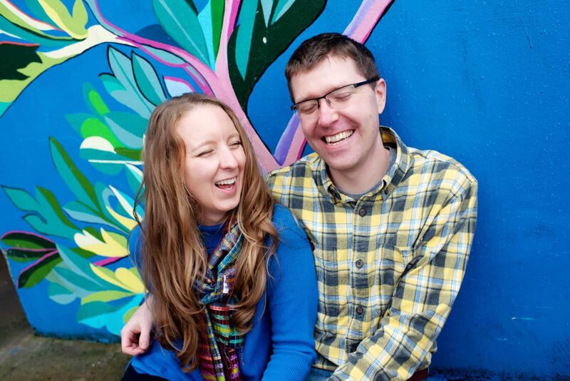 a man and woman sit and laugh with closed eyes in front of a colorful painted mural wall