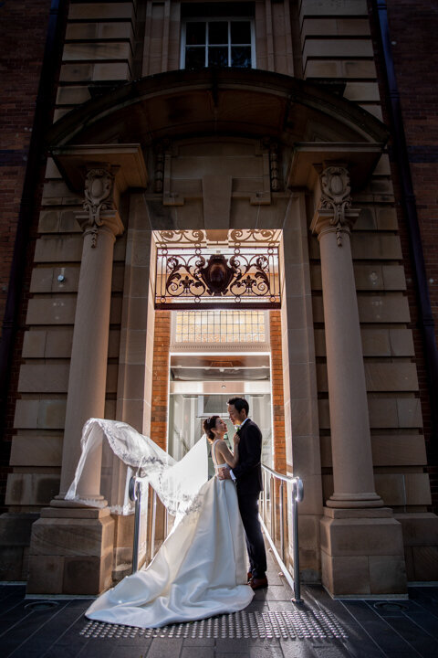 Bride and groom poses in front of the doorway with the brides flying veil