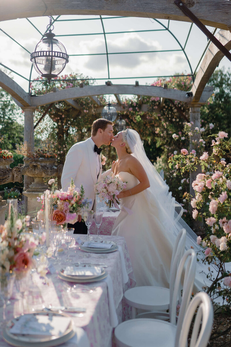 bride and groom kiss in a rose garden at euridge manor next to their wedding table decorated in pink patterned linen