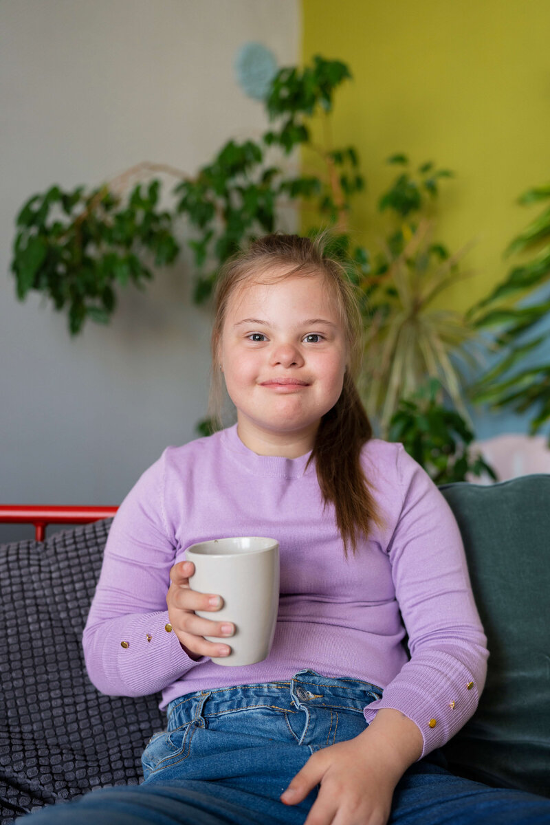 A young girl with Down syndrome, wearing a lavender sweater and jeans, is sitting on a couch holding a white mug, smiling softly with a background of green indoor plants.