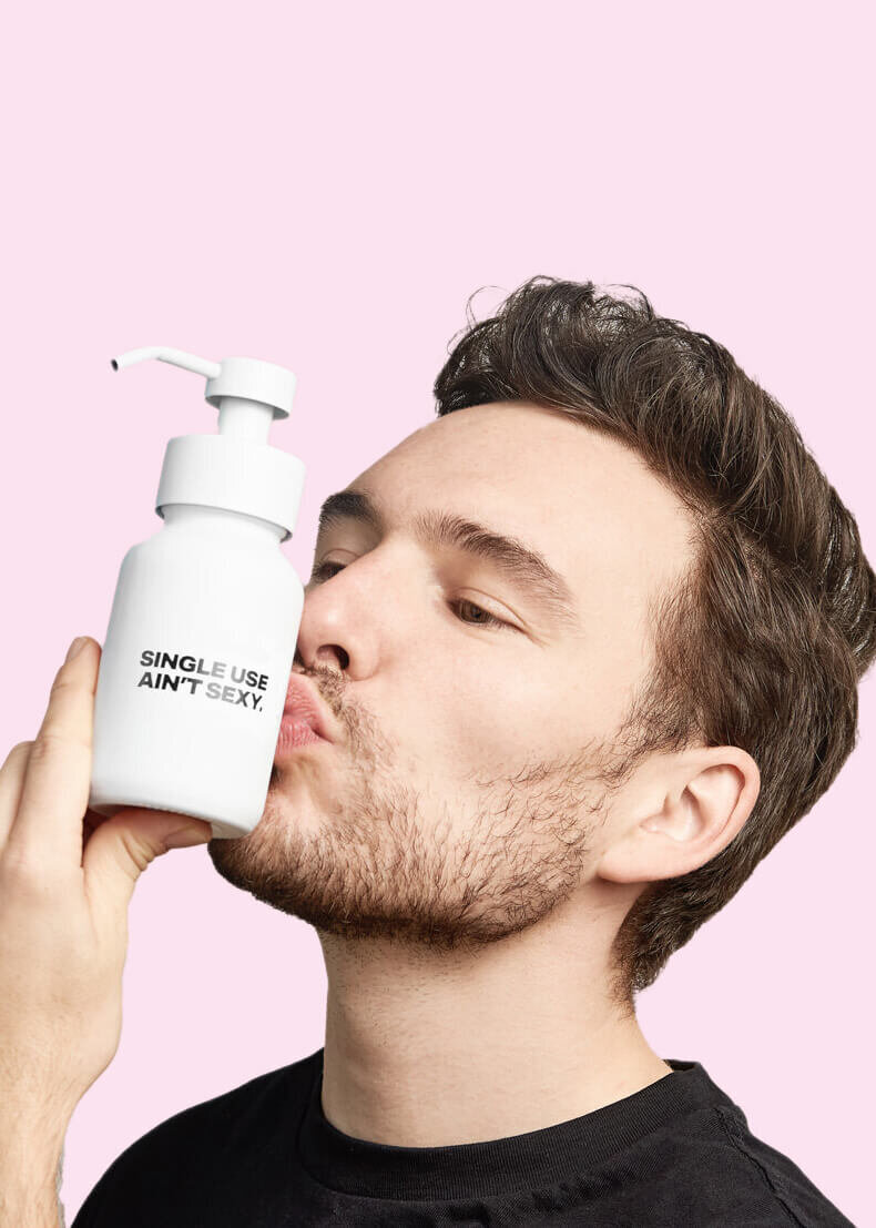 Handsome man kissing a Single Use Aint Sexy refillable hand-soap container against a pale pink background.