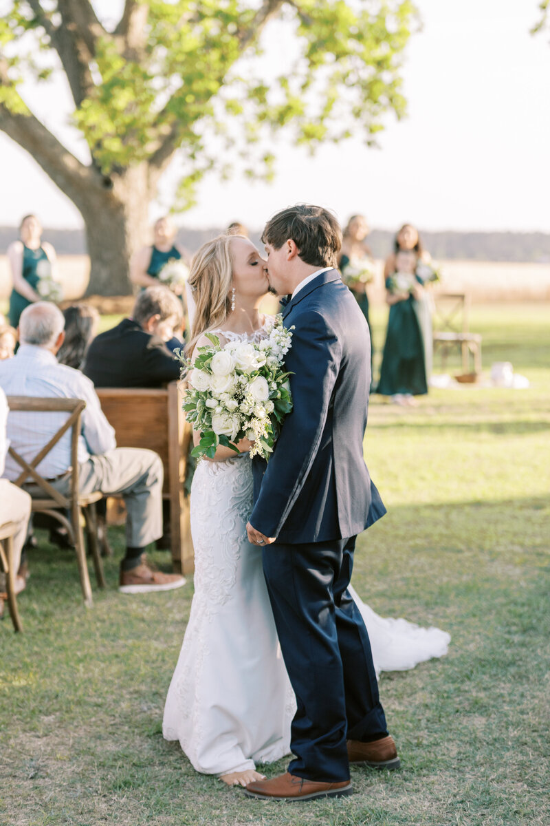 Groom in navy suit kisses bride in lace top dress holding bouquet at the end of the aisle after recessional. Aisle is flanked with oak stained chairs and pews with large oak in the background.
