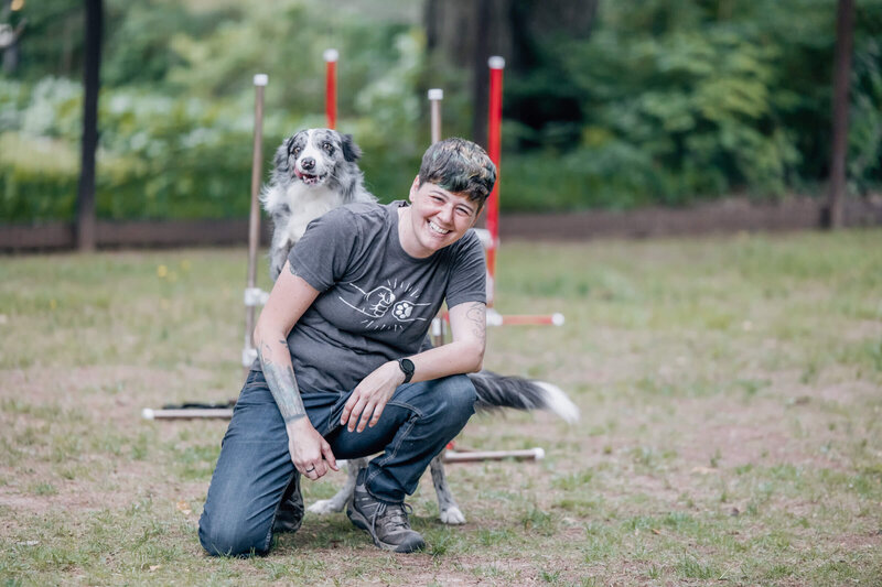 Dog trainer smiles at camera with grey and white dog peeking over her right shoulder.