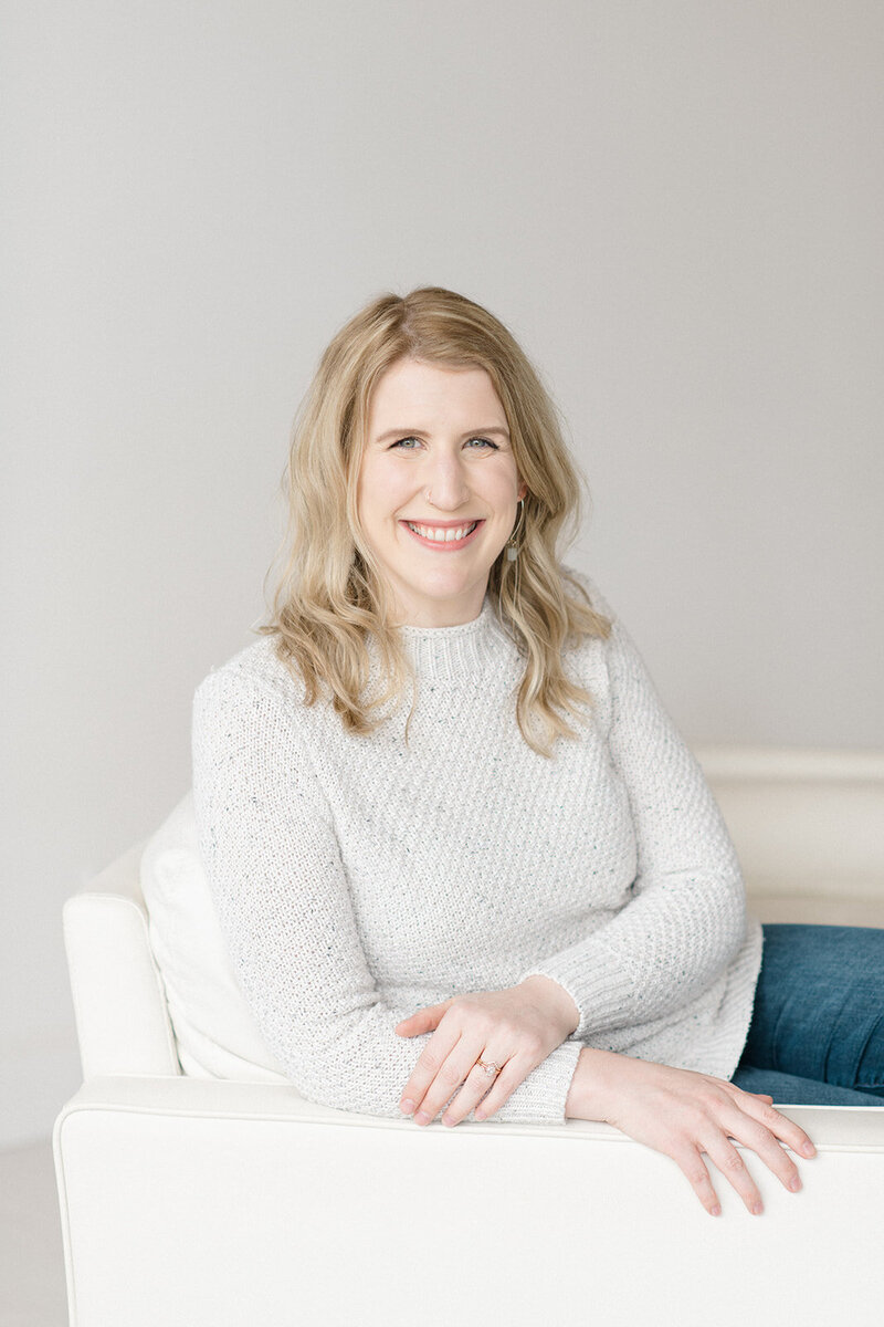 Photo of Shaunae Teske, a family photographer in Seattle, wearing a plaid blazer looking down at her camera and smiling while sitting on a stool