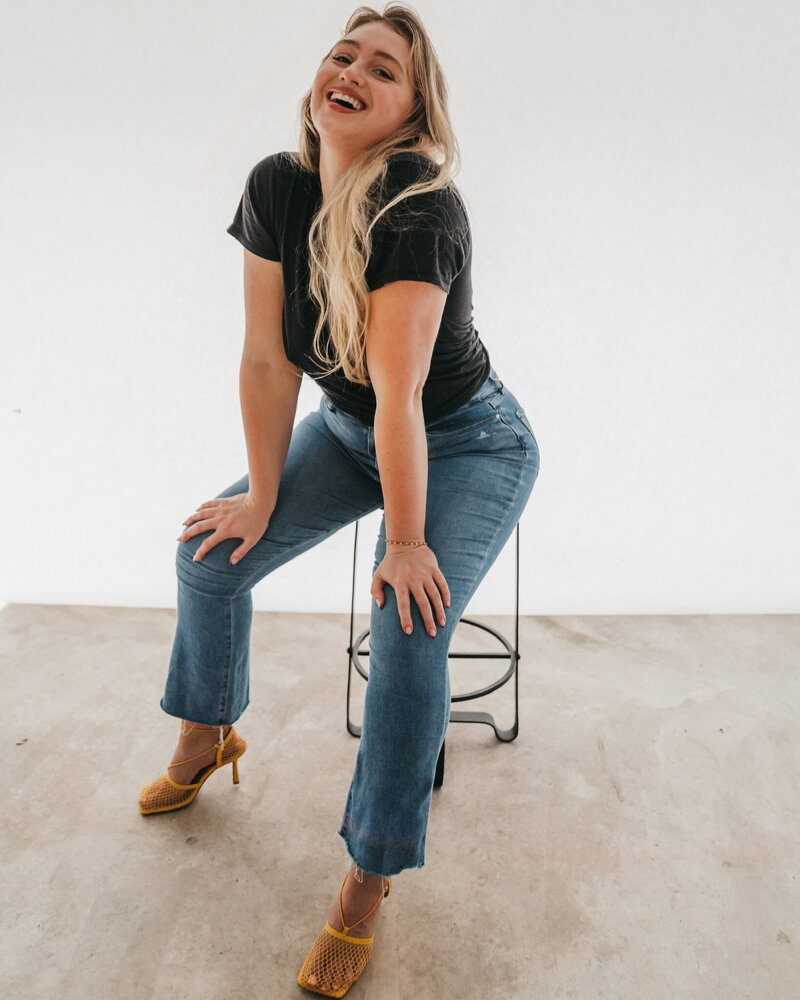 Iskra is wearing a casual tee with jeans, and she is posing on a stool, with her hands on her knees, and smiling into the camera.