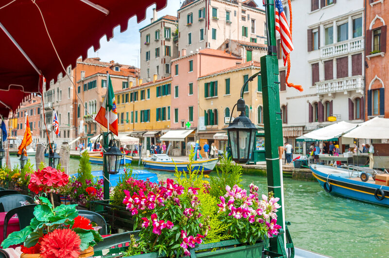 Colorful flowers adorn a railing overlooking a canal in Venice. Several boats float on the water, and a row of vibrant buildings lines the canal. Flags hang above, and people stroll along the walkway on a clear day.