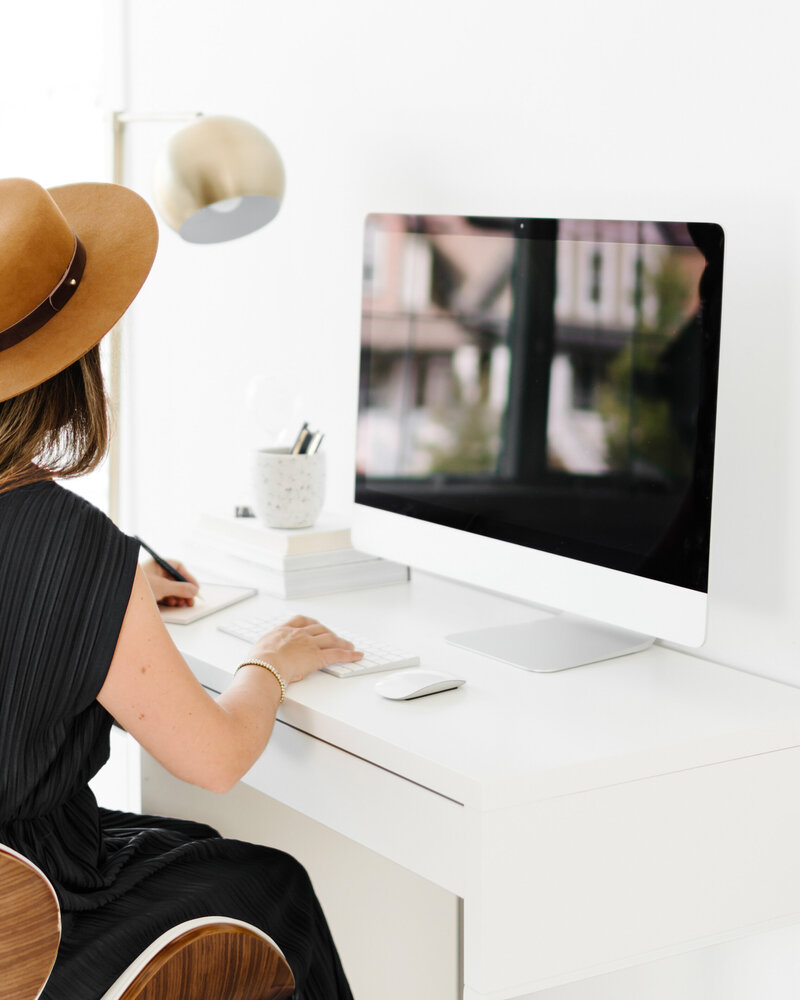 Woman in brown fedora sitting at a white desk with a large computer