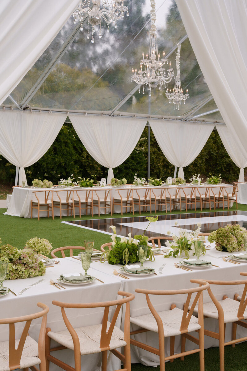 Tent reception setup with long tables, green and white florals, and chandeliers overhead.