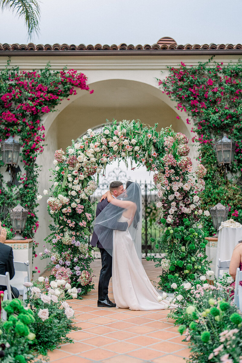 Bride and groom share their first kiss under a lush floral arch at their wedding ceremony at Hummingbird Nest Ranch in Simi Valley
