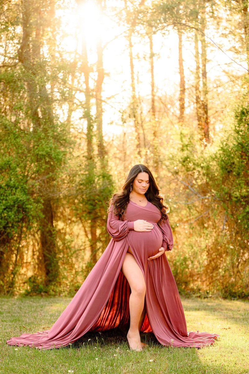 Pregnant mother is posed with both hands on her belly and her leg exposed due to the slit in her rose pink maternity dress looking down towards her belly in an outdoor setting