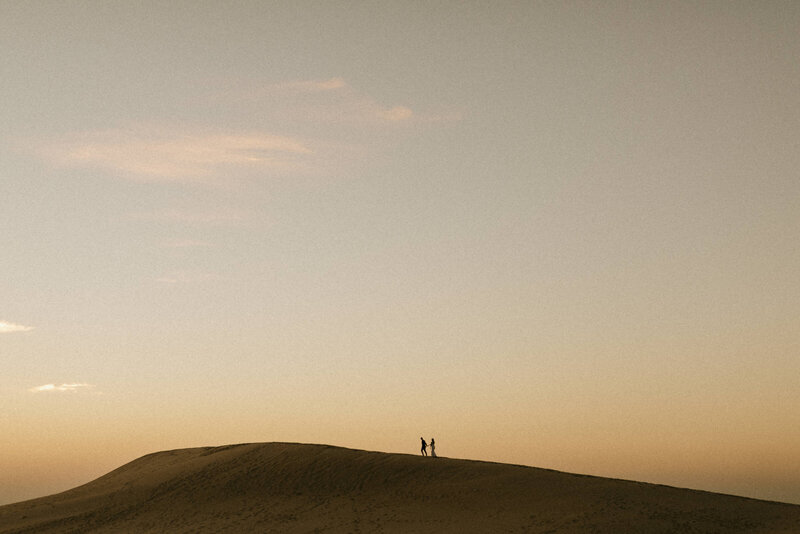 A couple holding hands and walking along the ridge of a sand dune in the distance